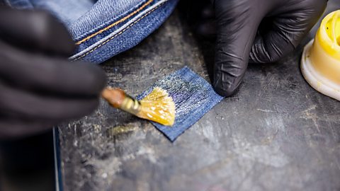 An adult hand with black gloves on, gluing a small square piece of denim with glue and a brush.