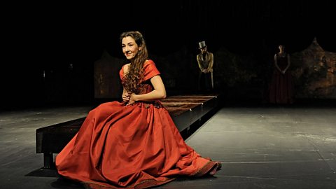 A lady in a red dress sits on a bench in a Spanish production of 'The Merchant of Venice'.