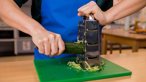 A close up of a person's hands, one holding the top of a grater on a green chopping board, while the other hand firmly grips a  cucumber and peels down the grater.