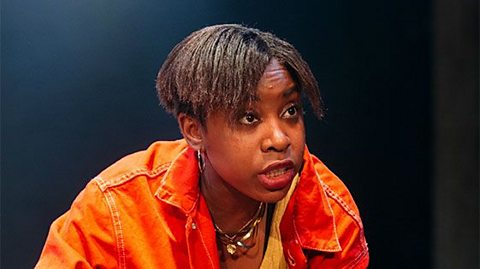 An actor playing Del. She is wearing a bright orange jacket and has short hair.