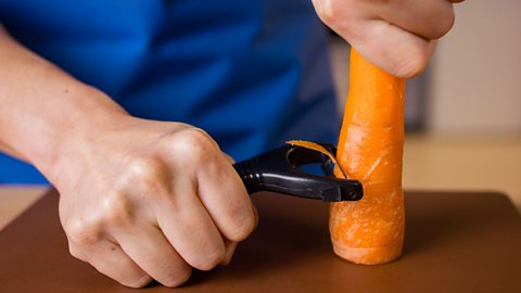 A close up of a person's hands, one holding a half peeled carrot on its end, while the other hand firmly grips a peeler and peels the carrot half way down.