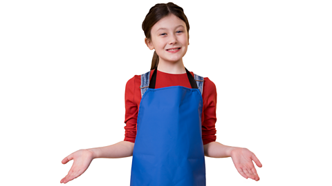 A young girl standing wearing a red top, jeans and trainers, with a blue apron on top. Her arms are out at her sides with palms open.