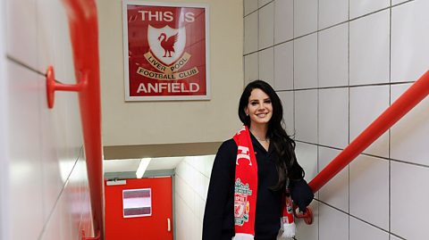 Lana Del Rey wearing a LIverpool FC scarf. A poster above her states, 'This is Anfield'. 