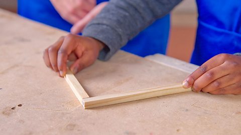 Hands holding two pieces of wood end to end at a 90 degree angle on a table