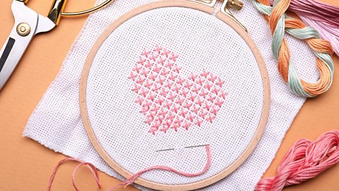 A cross stitched pink heart with different coloured thread and scissors lay around