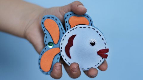 A child's hand holding a fish made from felt and decorated with googly eyes and sequins