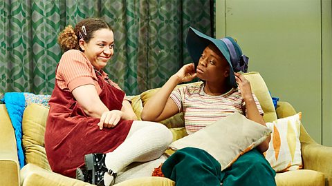 Two girls sitting on a yellow couch. One is wearing a red pinafore dress and the other is wearing green trousers with a stripey top.
