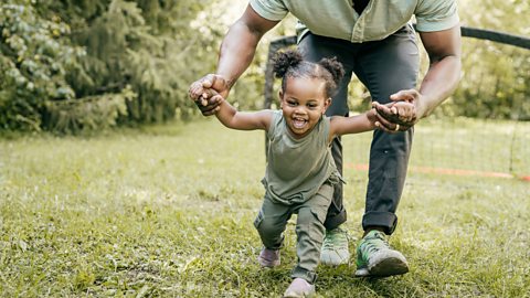 A young girl strides forward into the garden, huge grin on her face, supported by her father, who is helping her to walk so confidently.