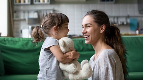 A mother and daughter share a moment, talking about feelings with a smile on each of their faces. The daughter holds a white teddy bear,