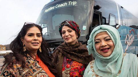 Three South Asian women wearing traditional clothing in front of a blue coach which says Aunties Coach Trip on the front display