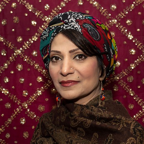 A South Asian women wearing dark red traditional clothing stands in front of a red and gold pattern backdrop