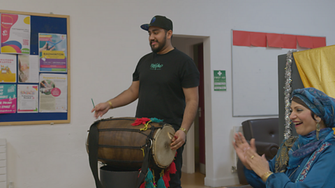 A South Asian man in his twenties plays a dhol drum, situated horizontally