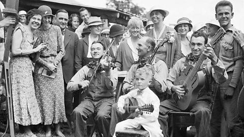 Four country musicians are sat down with guitars, while a crowd, including the First Lady Eleanor Roosevelt, are standing behind watching them.