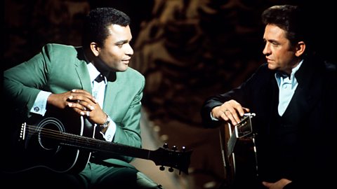 Charley Pride, wearing a green suit and leaning on a guitar, looks at Johnny Cash, dressed in a black suit and white shirt, also leaning on a guitar