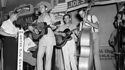 Hank Williams is front of stage, wearing a light suit and cowboy hat, playing guitar while singing. He is supported by several other musicians, playing guitars and double basses.