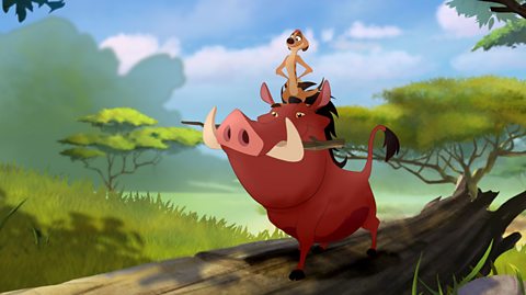 Timon, the meercat from the Lion King, is sat on the head of Pumbaa, a warthog. Pumbaa is stood on a log and has a stick in his mouth.