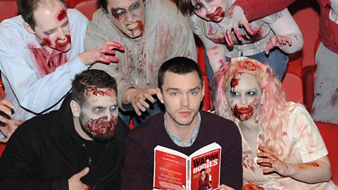 Nicholas Hoult is sat centrally, holding a copy of the book Warm Bodies. He is surrounded by actors wearing zombie makeup pretending to grab him.