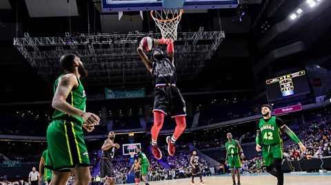 Bounce Moody, wearing a black and red Harlem Globetrotters kit, in mid-air, ready to dunk the ball into the basket, while several Washington Generals players, in green and black kit, watch on