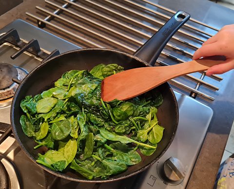 A frying pan full of spinach with a wooden spatula mixing.