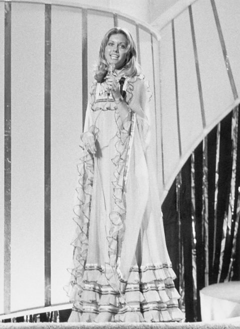 Black and white image of the singer Olivia Newton-John representing the UK at Eurovision 1974 in Brighton. She has long blonde hair and is wearing a floor-length dress with lots of frills and is singing into a microphone