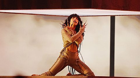 The Swedish singer Loreen performing her winning son Tattoo at the 2023 Eurovision Song Contest. She has long, dark curly hair, long nails and is wearing a beige and brown bodysuit while singing beneath an illuminated box