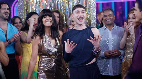 The singer Olly Alexander surrounded by the stars of Strictly Come Dancing as he announced his participation in Eurovision 2024 for the UK. He has short, cropped, dark hair and is wearing an off-the-shoulder top with black pants and is stood next to a smiling Claudia Winkleman