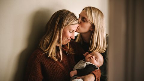 A young couple cradle their baby - one mother kissing the other on the forehead.