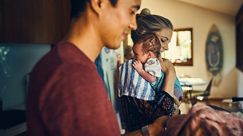 A mother cradles her baby over her shoulder while her husband works in the kitchen.