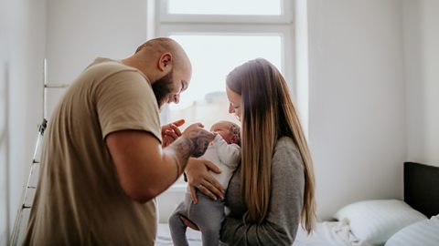 A couple fuss over their baby - the mum cradling them, the dad smiling and holding their hands. The bedroom in the background looks newly decorated,