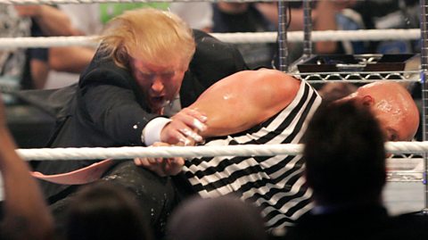 Donald Trump falls down to the ground, having been hit with a Stone Cold Stunner wrestling move by Stone Cold Steve Austin. Austin is wearing a white and black striped sleeveless shirt while Trump is in a dark suit.