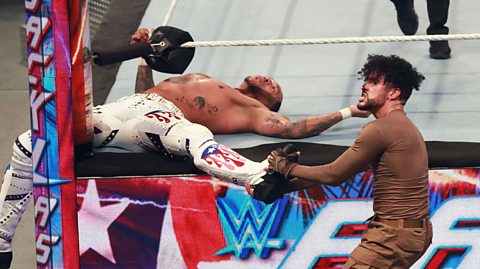 Bad Bunny is to the right, wearing all brown, outside the ring. He is holding the boot of Damian Priest, who is wearing white wrestling tights and boots and is on his back in the ring, with his legs either side of the ring post. Bad Bunny is set to drive Priest's leg into the post.