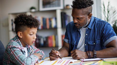 Father helping son with homework at table with books open in front of them.