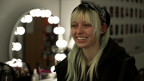 Junior hair and make-up artist, Cat, sat smiling in front of a make-up mirror. 