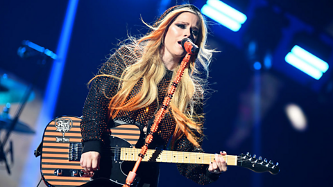 An image of Avril Lavigne holding a guitar in front of a microphone.