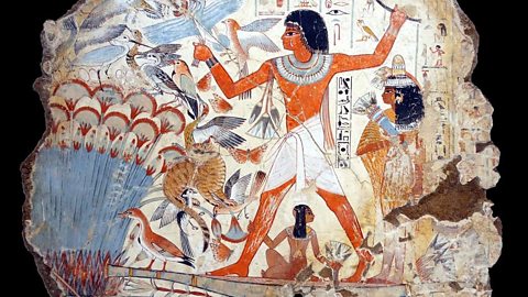 An Ancient Egyptian painting with a male figure wearing a white cloth around his waist, stands on a flat boat, spear in hand. Also on the boat are a dark-haired woman and child. Fish can be seen in the water, birds in the sky and a cat among the reeds. 