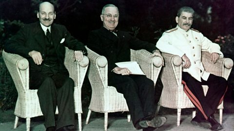 Three men sit in wicker chairs, two wearing dark suits (Attlee and Truman) and one wearing a white suit jacket with black and red trousers (Stalin)