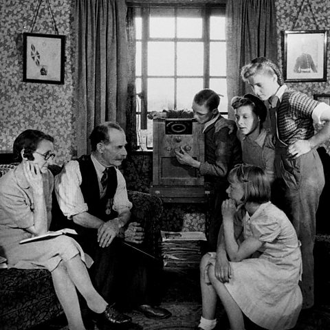 Radio was a major medium of communication in 1945, this image shows a multi-generational British family of six gathered around a wireless in the 1940s, listening intently to a broadcast 