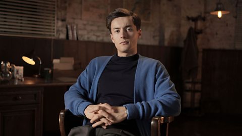 Actor Andre Frey, a young white man with dark brown hair and green-brown eyes, portrays French resistance member André Heintz. He is sat in a chair, wearing a black top with a blue cardigan, and is against an industrial sand-brick background.