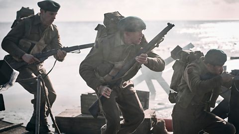 Several young male actors in British uniforms run along a beach with guns in hand.