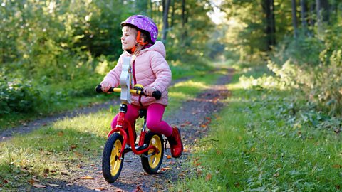 A pre-school girl in a pink coat and a purple helmet rides a balance bike through some woods.