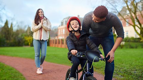 Dad helps his son balance on his bike on a path, while Mum looks on