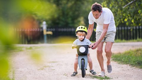 A pre-school boy in a yellow helmet sits on a balance bike, supported by his grandfather.