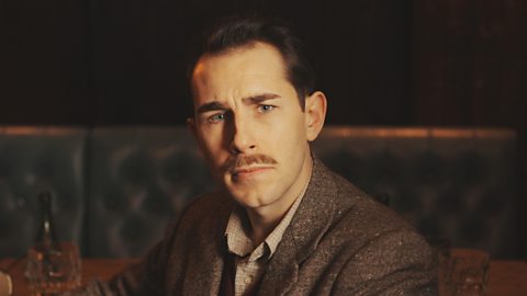 Actor Samuel Lawrence playing Private Wally Parr, a white man with brown hair and a thin moustache. He is dressed in brown courdroy and sat in a dimly-lit cafe.