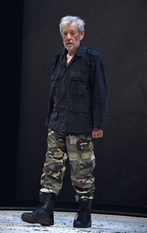 An older man with white hair and beard, wearing black and camouflage combat fatigues, stands in the middle of a stage