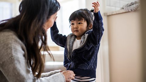 A mother puts a cardigan on her daughter, whose arms are in the air.