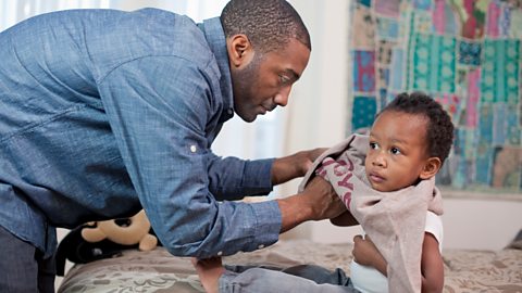 A dad carefully pulls a jumper onto his young child.