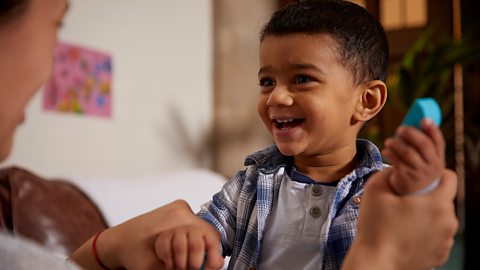 A young boy smiles while playing with his mum, holding a bright blue building block.