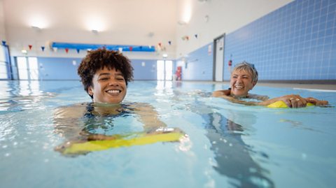 Two women in a community pool, smiling and holding out yellow floating boards. The left is a middle-aged light-skinned black woman with short curly hair, and the right is an older white woman with very short grey hair.