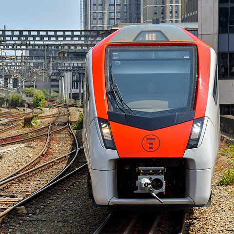 A red and grey train travelling on a train track with a city landscape in the background.