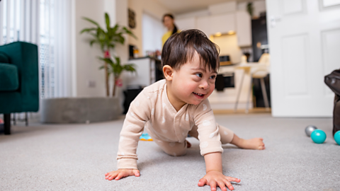 A baby with Down's syndrome crawls across a carpeted floor. A parent watches on from the background.